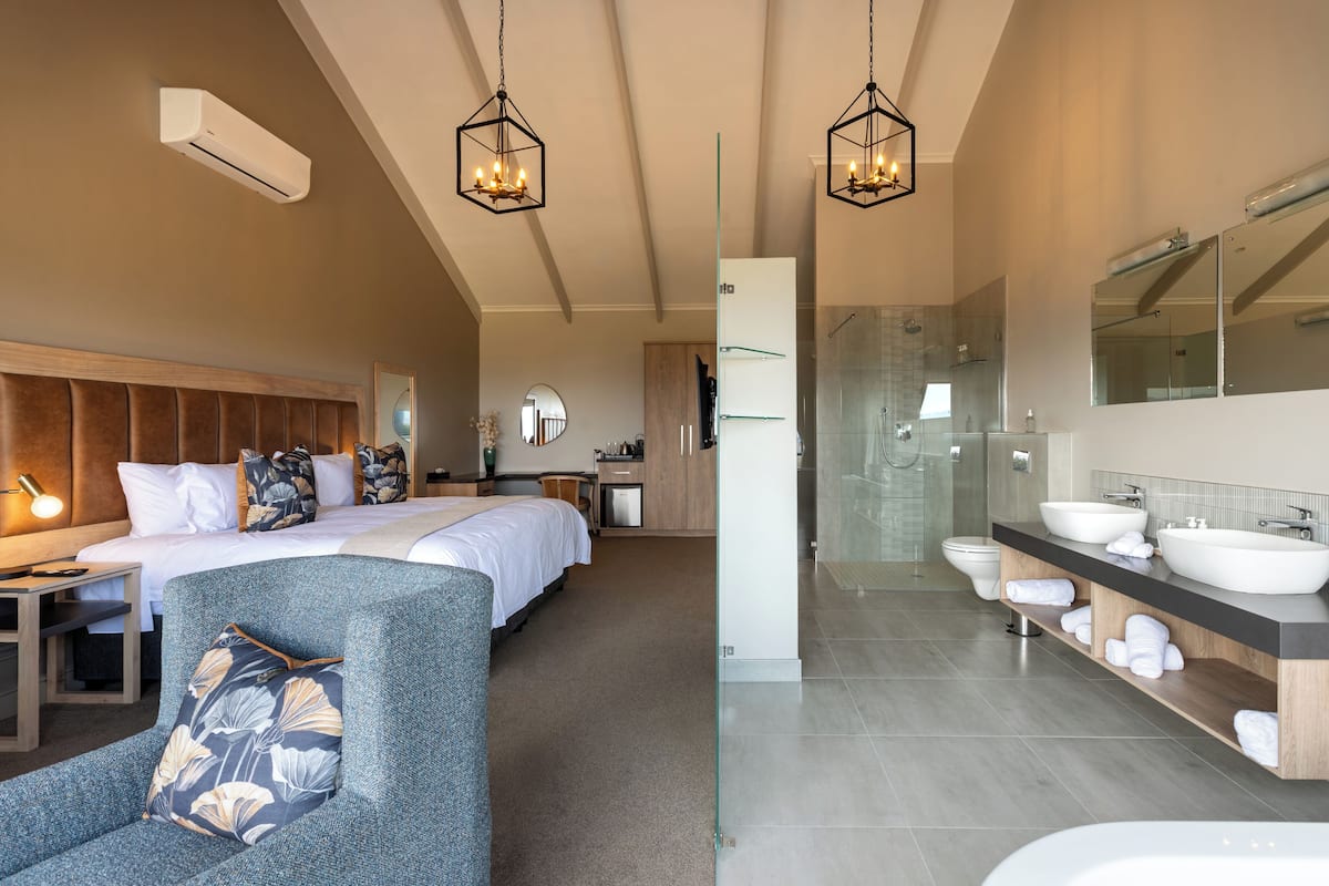 A modern hotel room with a large bed, a sitting area, a bathroom with a large mirror, and a shower.