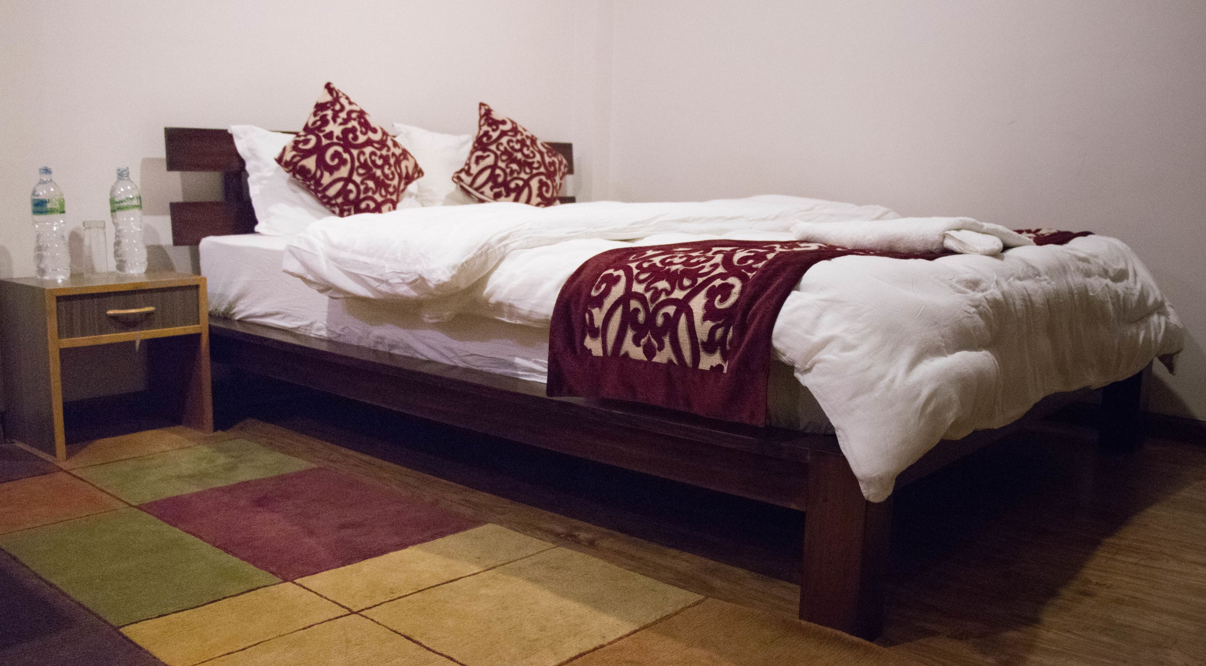 A neatly made bed with a patterned throw, a bedside table with bottles, and a colorful rug.