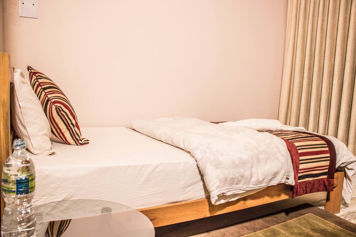 Uma cama simples com estrutura de madeira, colchão branco e manta listrada. Há uma garrafa de água sobre uma mesinha ao lado da cama.