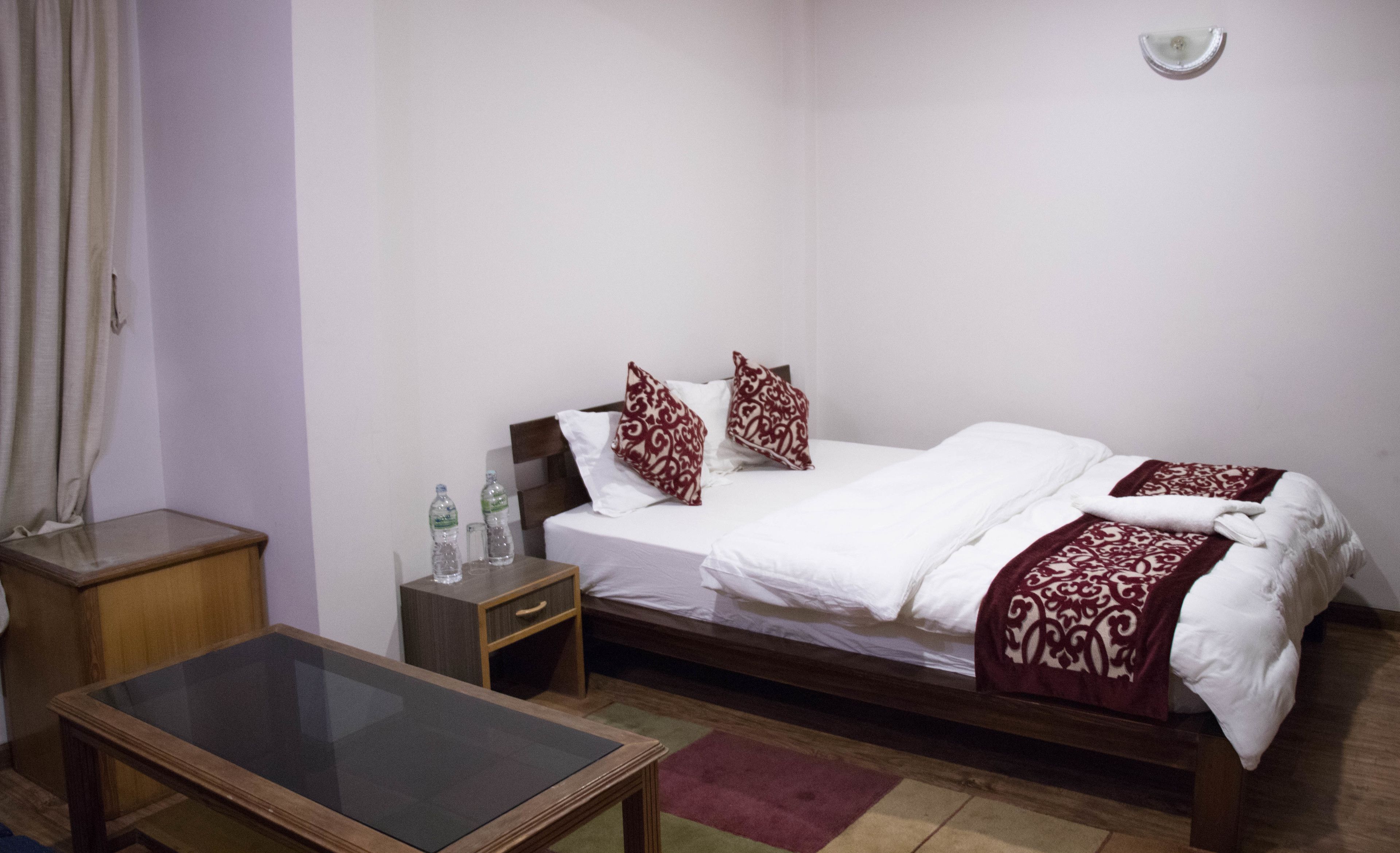 A hotel room with a single bed, two pillows, a bedside table with a glass, and a small table with a glass.
