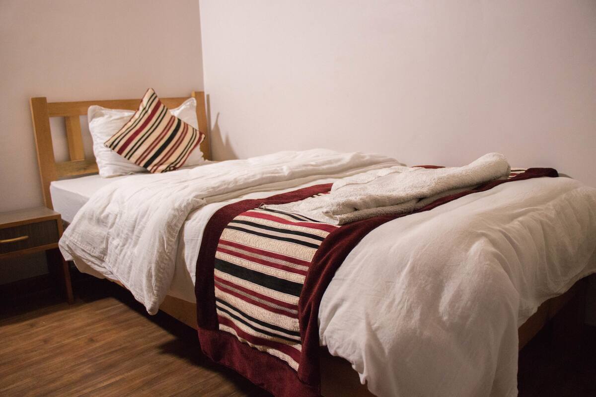 Uma cama de solteiro com cabeceira de madeira, manta listrada e travesseiro branco.