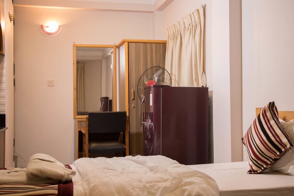 Quarto de hotel com cama, escrivaninha, cadeira, guarda-roupa, espelho e ventoinha.