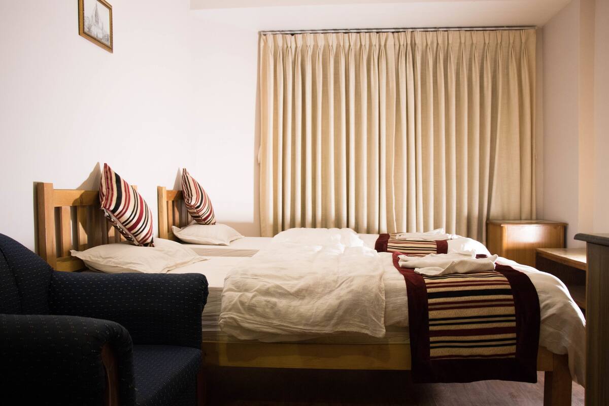 Quarto de hotel com duas camas, uma cadeira, uma mesa de cabeceira e uma cortina.