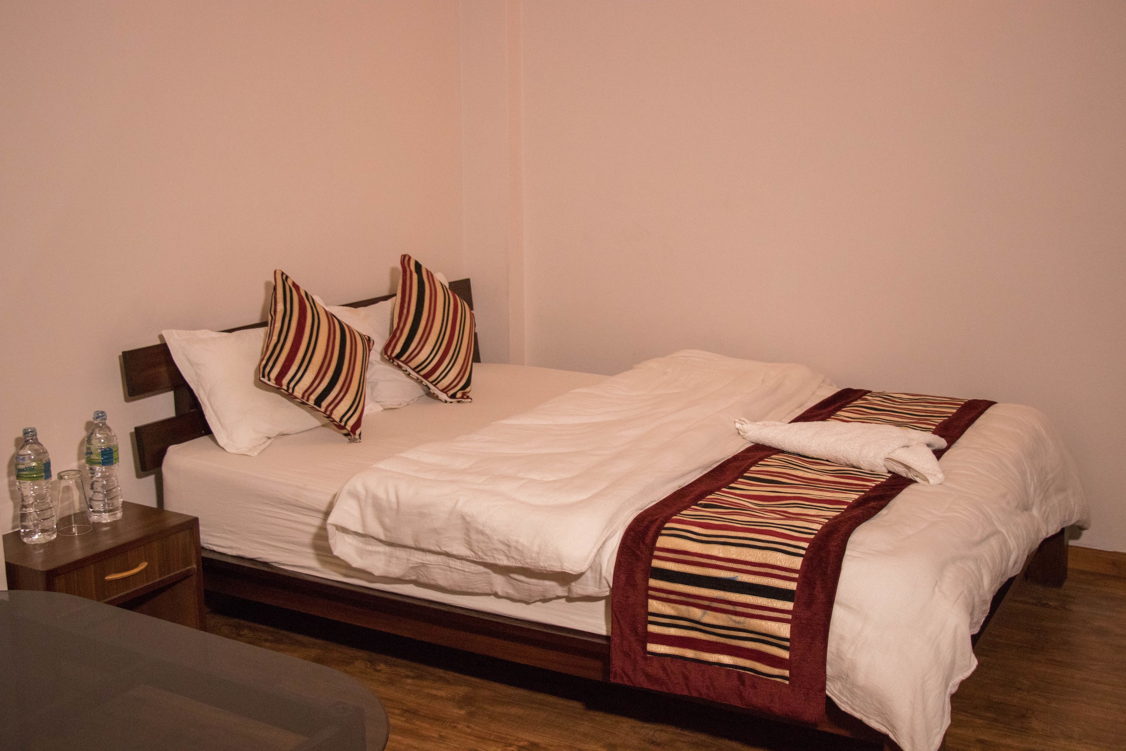 Una cama bien hecha con sábanas blancas y almohadas a rayas, una mesita de noche de madera con botellas de agua y un suelo de madera.