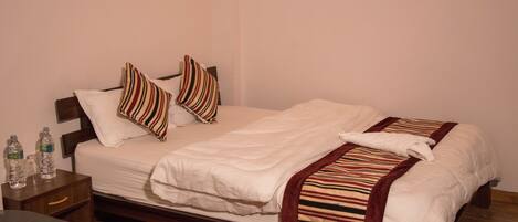 Una cama bien hecha con sábanas blancas y almohadas a rayas, una mesita de noche de madera con botellas de agua y un suelo de madera.