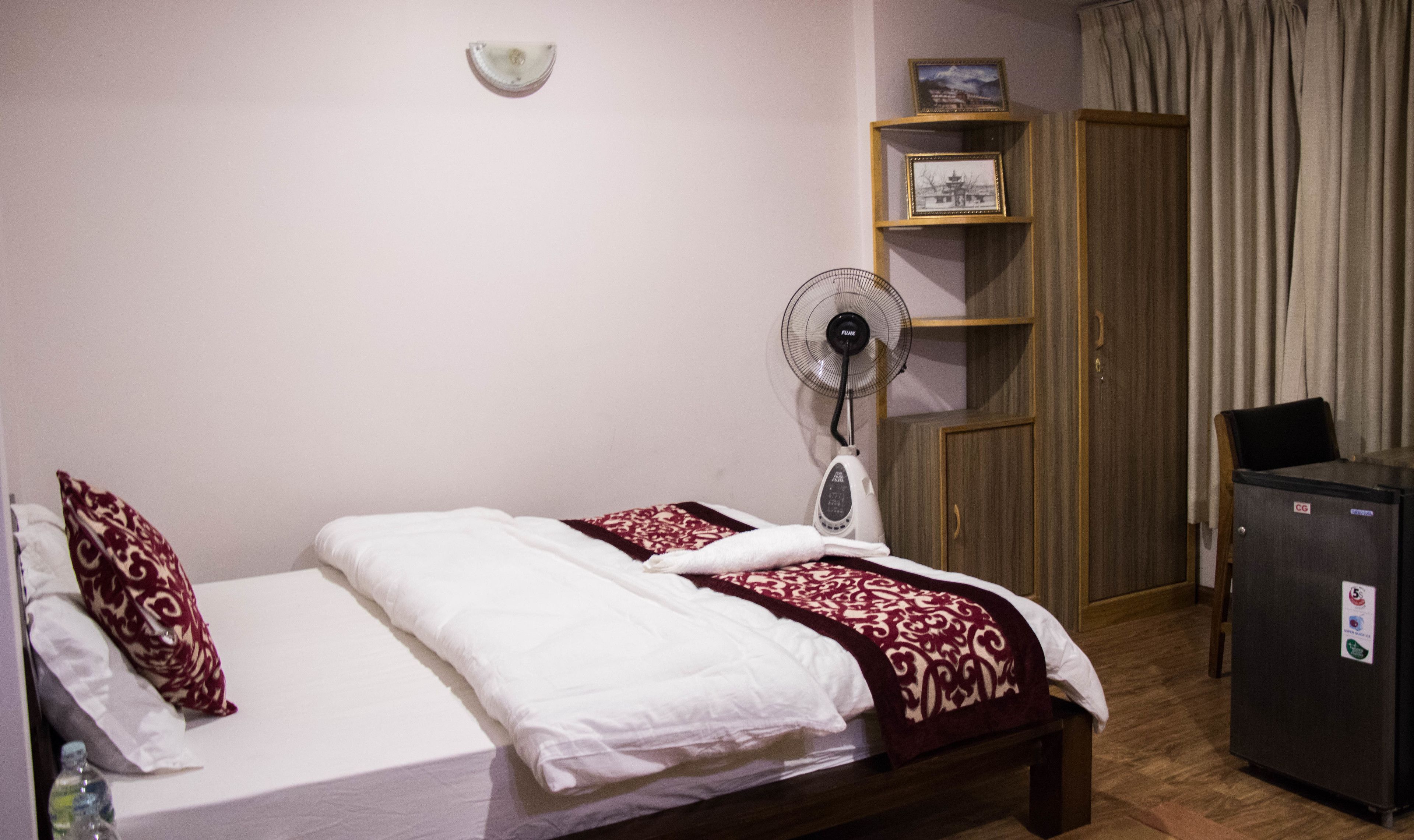 A single bed with white bedding and a red and white patterned throw, a standing fan, a wooden wardrobe, and a small refrigerator.