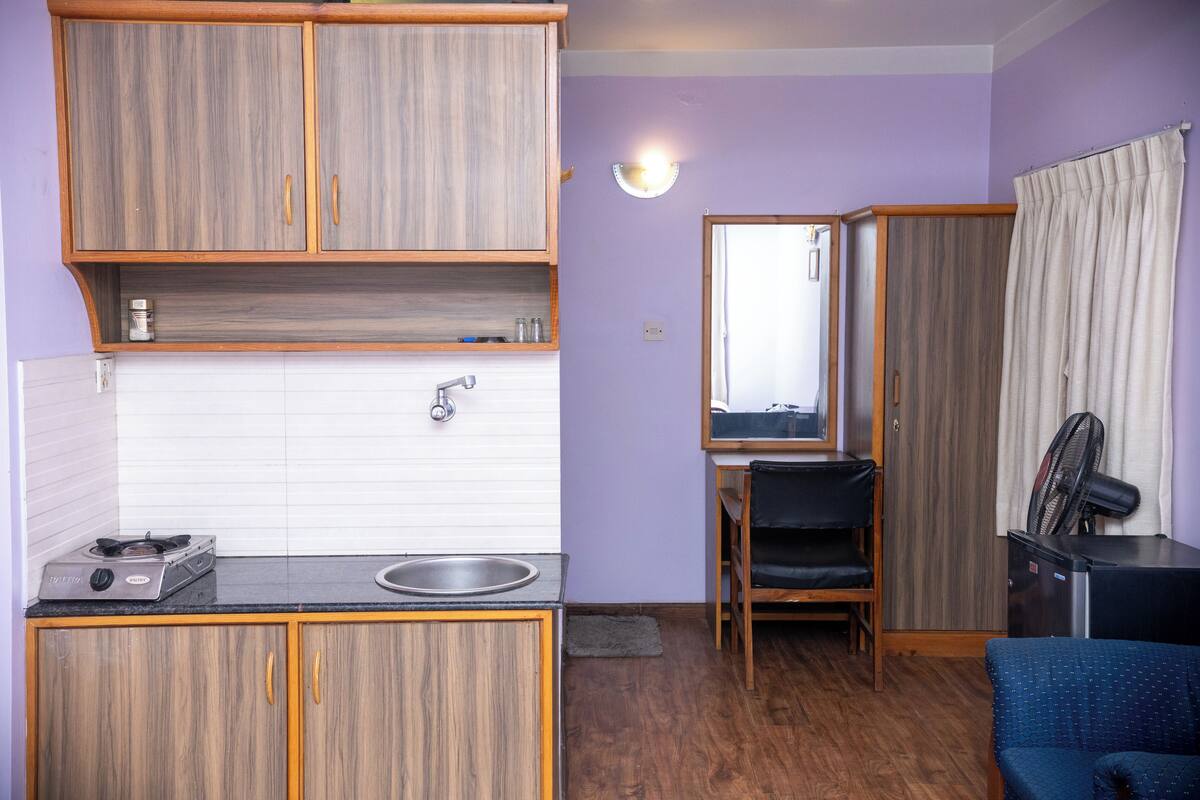Cozinha compacta com fogão, pia e armários de madeira. Há uma área de refeições com uma cadeira e uma mesa. Também se vê um espelho e um ventilador.