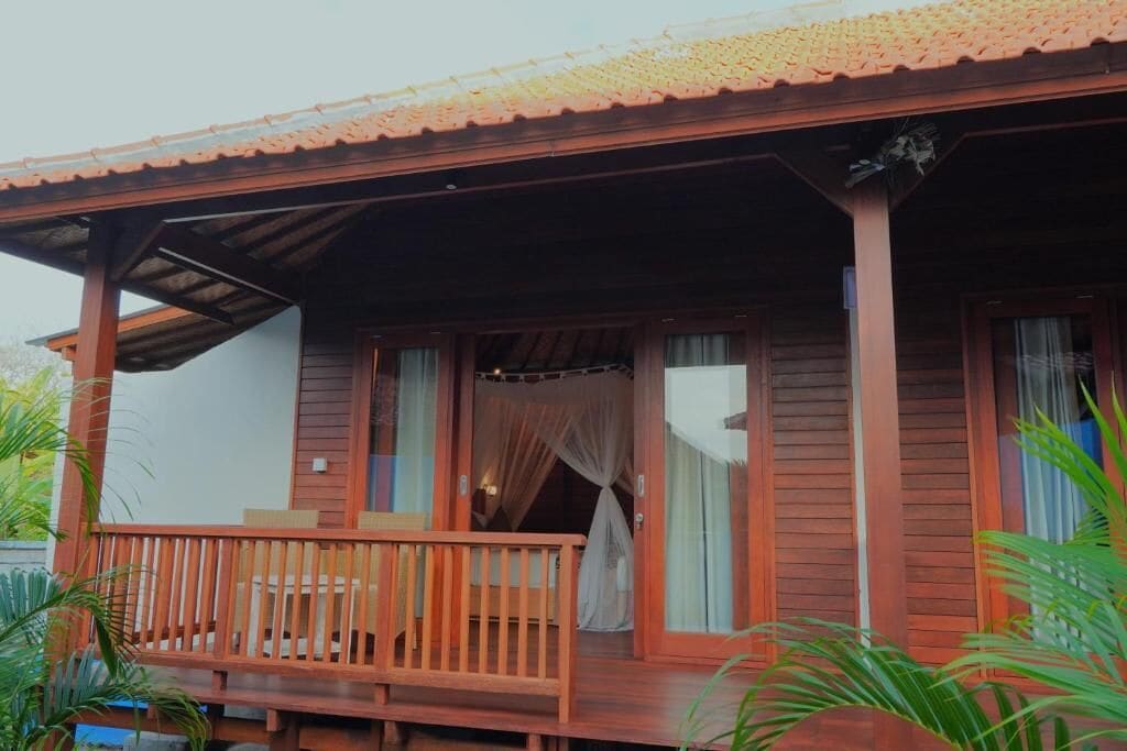 A wooden cabin with a balcony, a covered porch, and a window with a curtain.