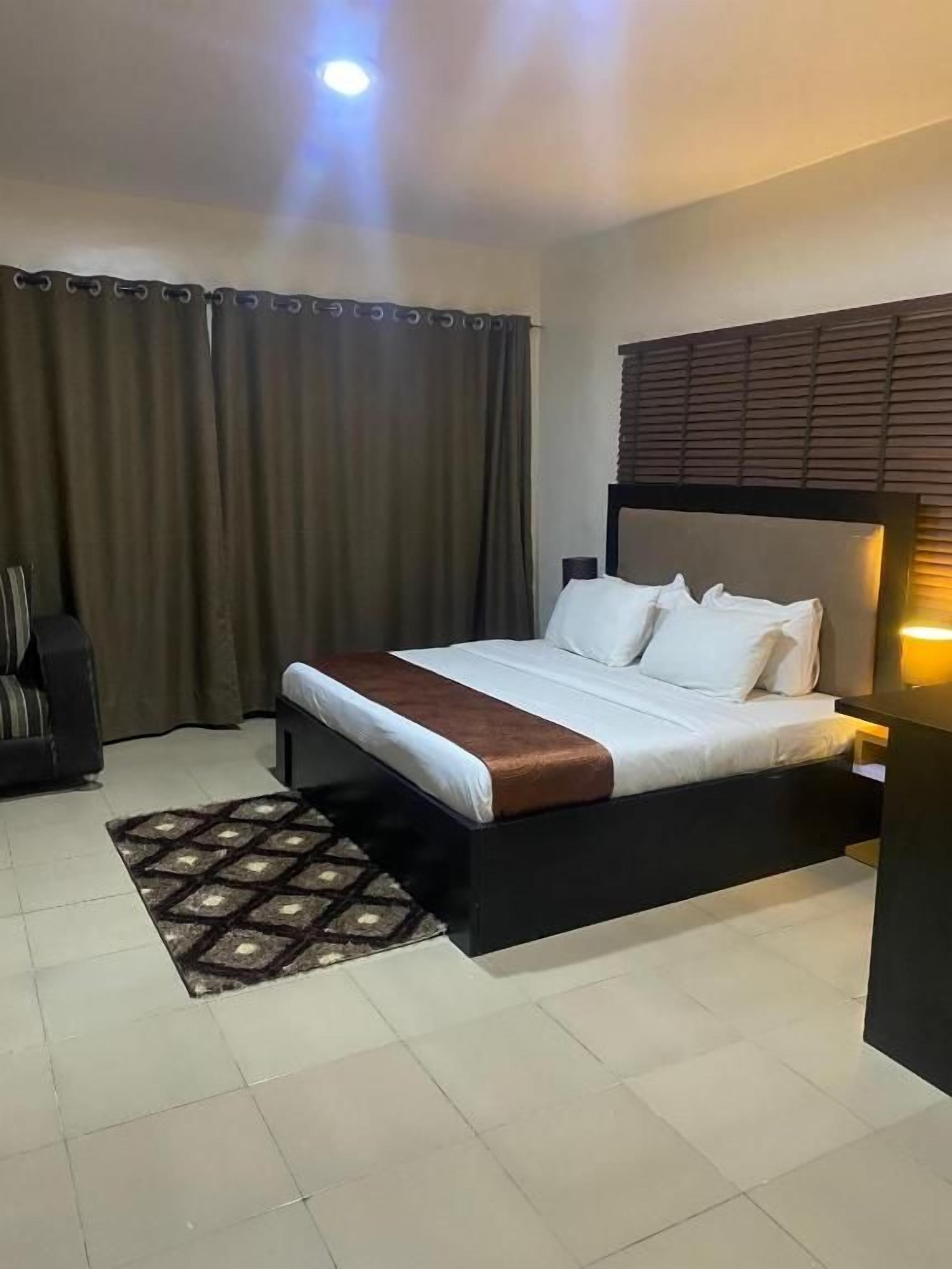 A hotel room with a bed, a nightstand, a lamp, a window with curtains, and a patterned rug.