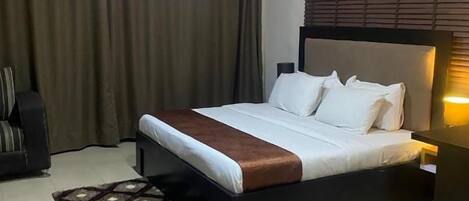 Una habitación de hotel con cama, mesita de noche, lámpara, ventana con cortinas y alfombra estampada.