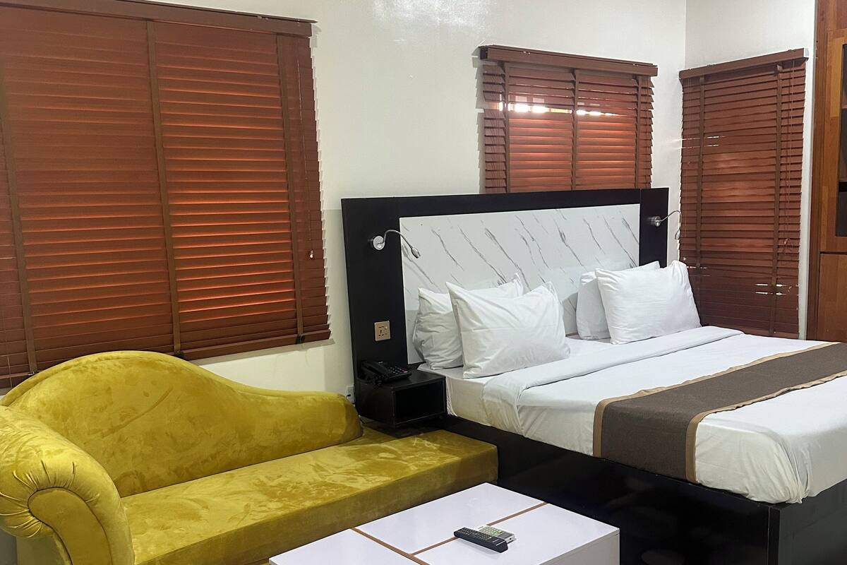 A hotel room with a bed, a yellow sofa, a white coffee table, and a patterned rug.