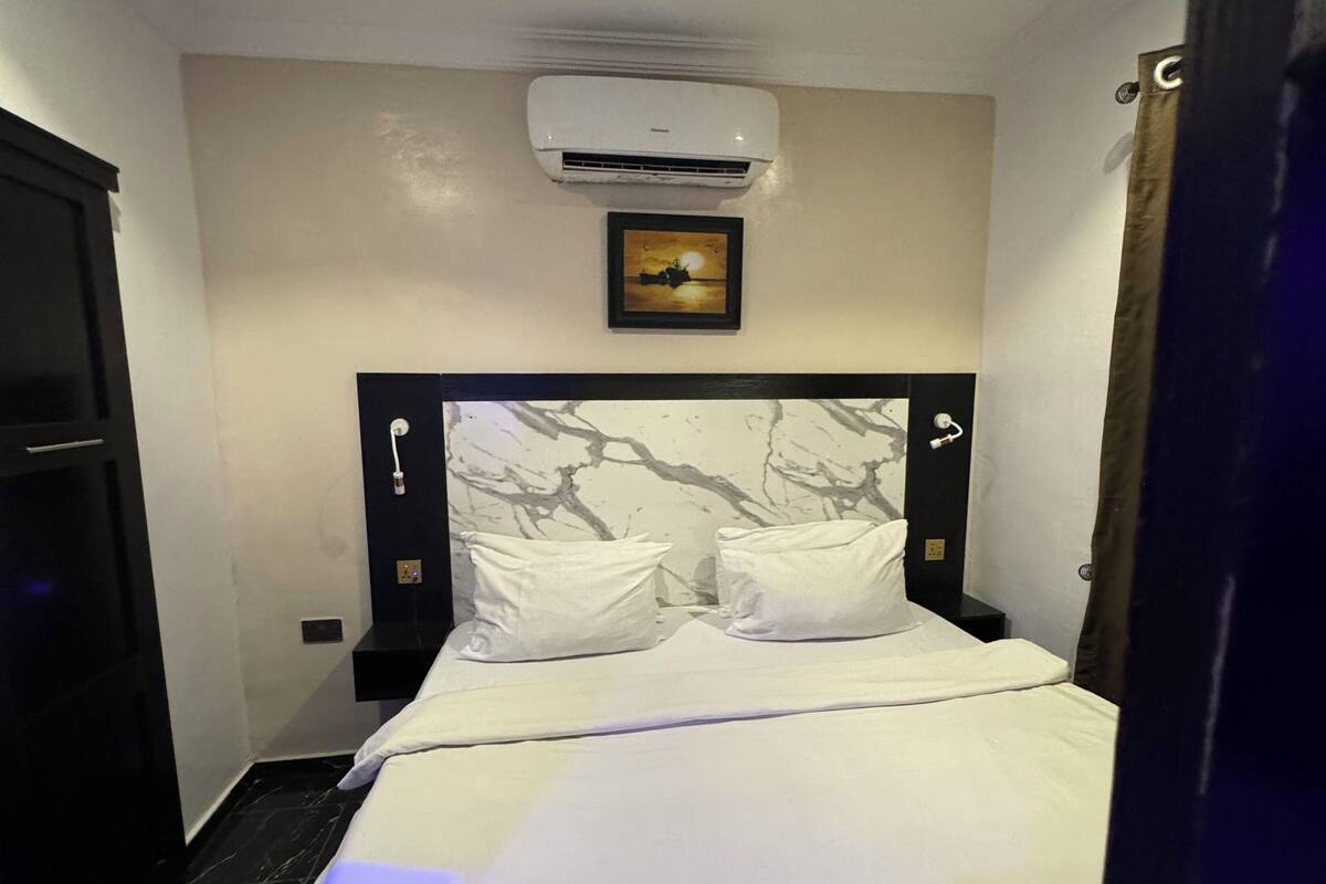 A hotel room with a bed, a wall-mounted air conditioner, a framed picture, and a marble-patterned headboard.