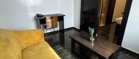 A room with a yellow sofa, a flat-screen TV mounted on the wall, a small wooden side table, and a dark wooden coffee table with a plant.