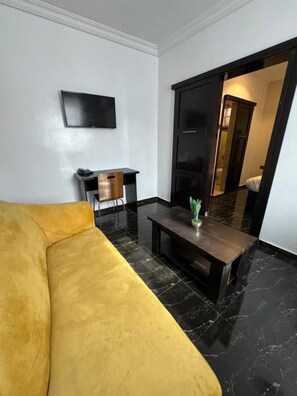 A room with a yellow sofa, a flat-screen TV mounted on the wall, a small wooden side table, and a dark wooden coffee table with a plant. - Posh Apartments Metro (Lagos)