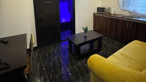 A compact living space with a black tiled floor, a yellow sofa, a small table with a plant, a kitchen area with a microwave, and a sliding door with blue lighting. - Posh Apartments Metro (Lagos)