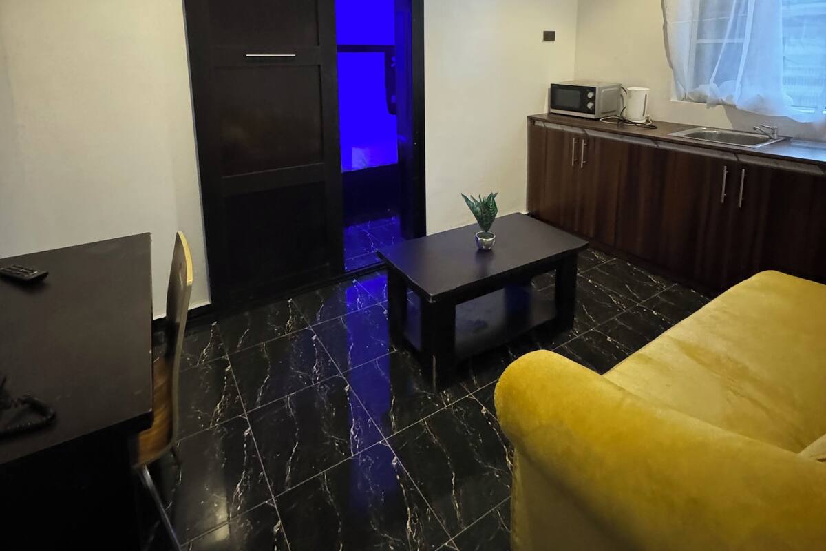 A compact living space with a black tiled floor, a yellow sofa, a small table with a plant, a kitchen area with a microwave, and a sliding door with blue lighting.