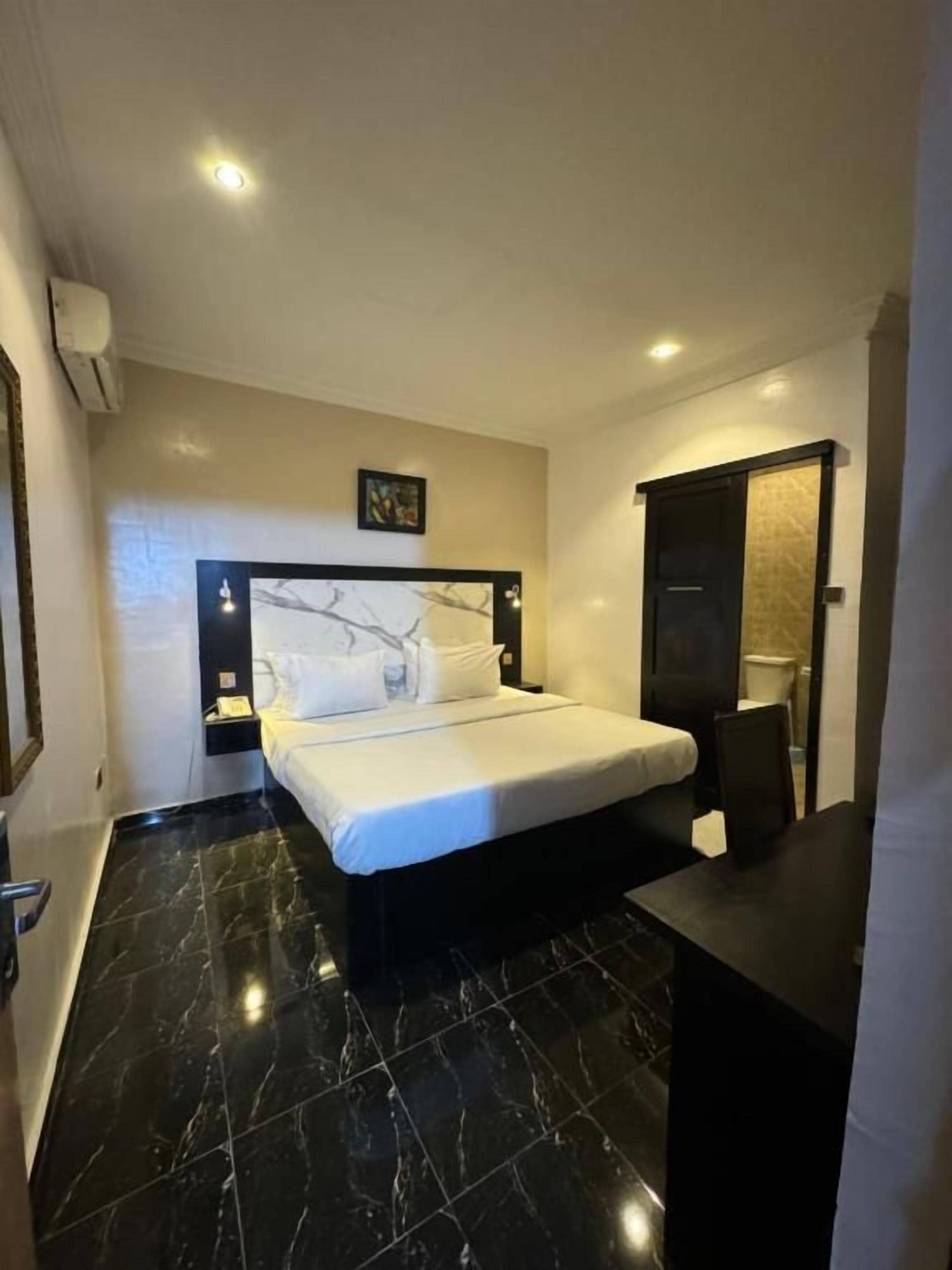 A hotel room with a bed, a bedside table, a black tiled floor, and a wall-mounted air conditioning unit.