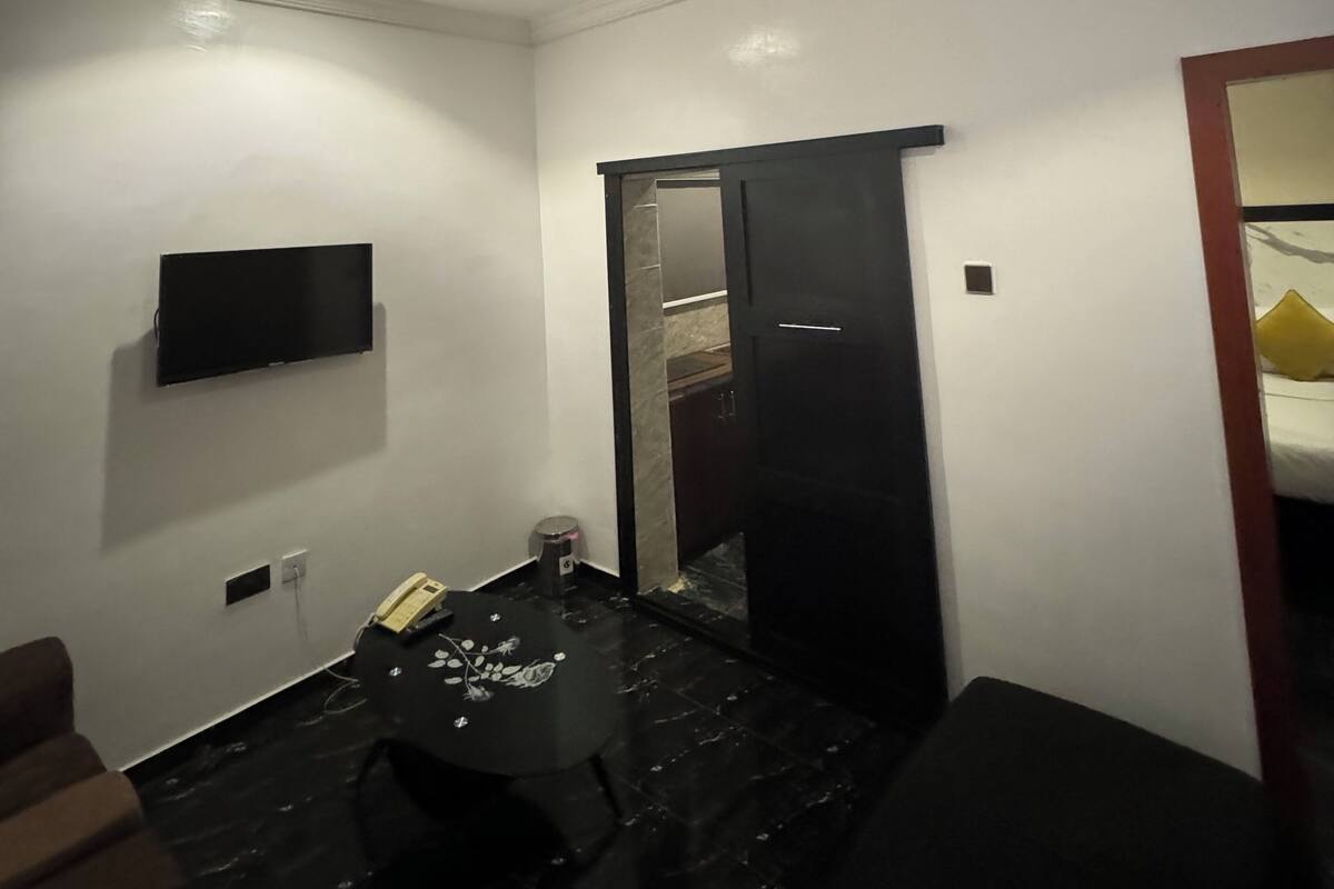 A small, well-lit room with a flat-screen TV mounted on the wall, a small table with a telephone, and a door leading to a bathroom.