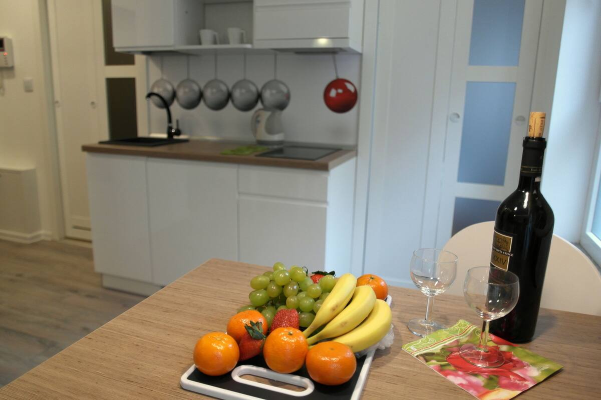 Une cuisine avec des armoires blanches, un plan de travail en bois et un plateau de fruits contenant des raisins, des bananes et des oranges. On y trouve également une bouteille de vin et deux verres sur le plan de travail.