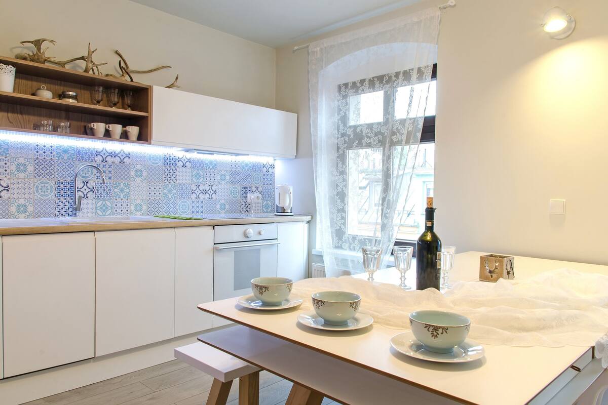 Une cuisine avec un plan de travail blanc, une table à manger avec des bols, une bouteille de vin et une fenêtre avec des rideaux.