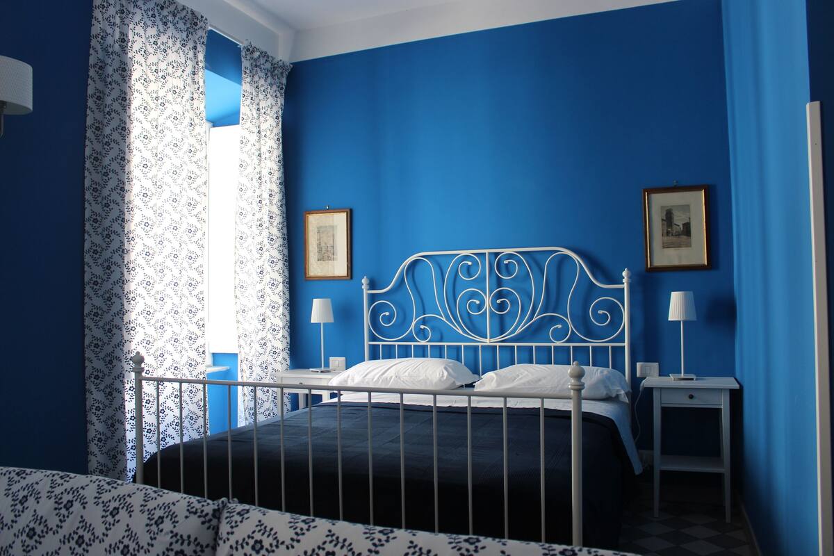 A bedroom with a blue wall, a white metal bed frame, two bedside tables with lamps, and a window with patterned curtains.