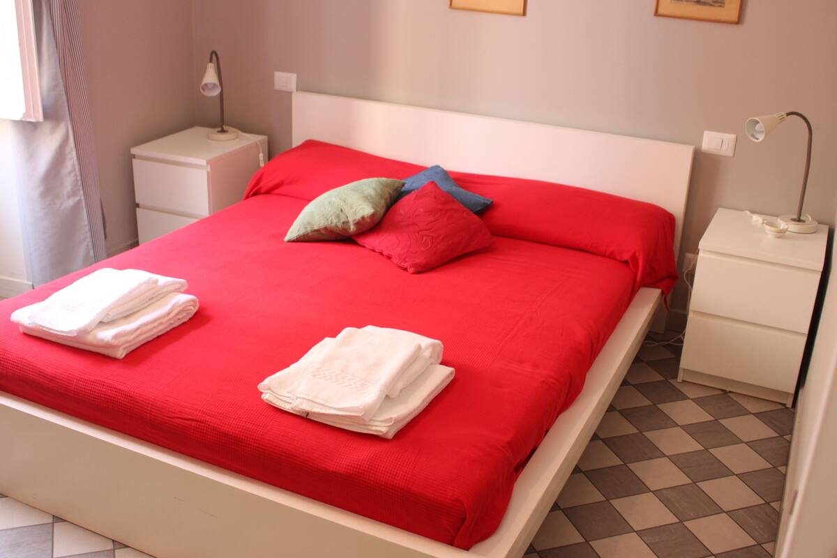 A neatly made bed with red bedding and white towels, two bedside tables with lamps, and a checkered floor.