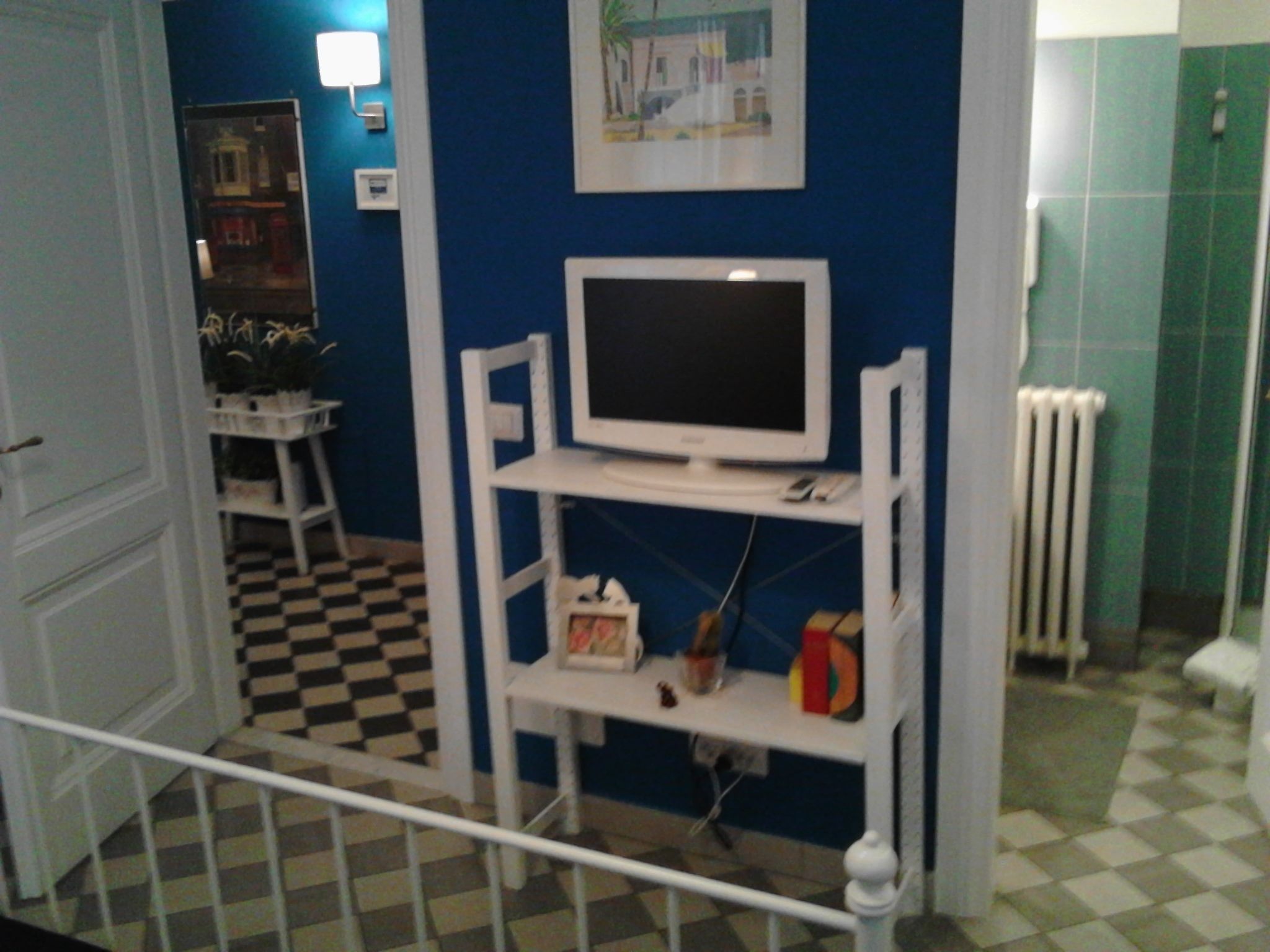 Una habitación con suelo a cuadros, un televisor sobre un pedestal blanco, un radiador y una puerta que da a otra estancia.