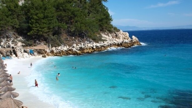 A beach with clear turquoise water, rocky coastline, and pine trees.