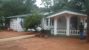 A small, single-story building with a green roof and white walls. There is a covered porch with a white railing and a person standing at the entrance.