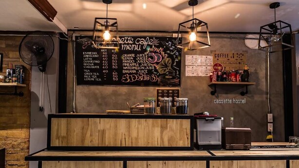 Una cafetería con un mostrador de madera, una pizarra con un menú que ofrece diversas bebidas y un estante con productos.