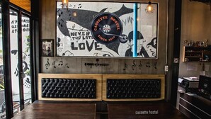 Interior de una cafetería con un cartel en la pared, dos zonas de estar con cojines y una barra de madera.
