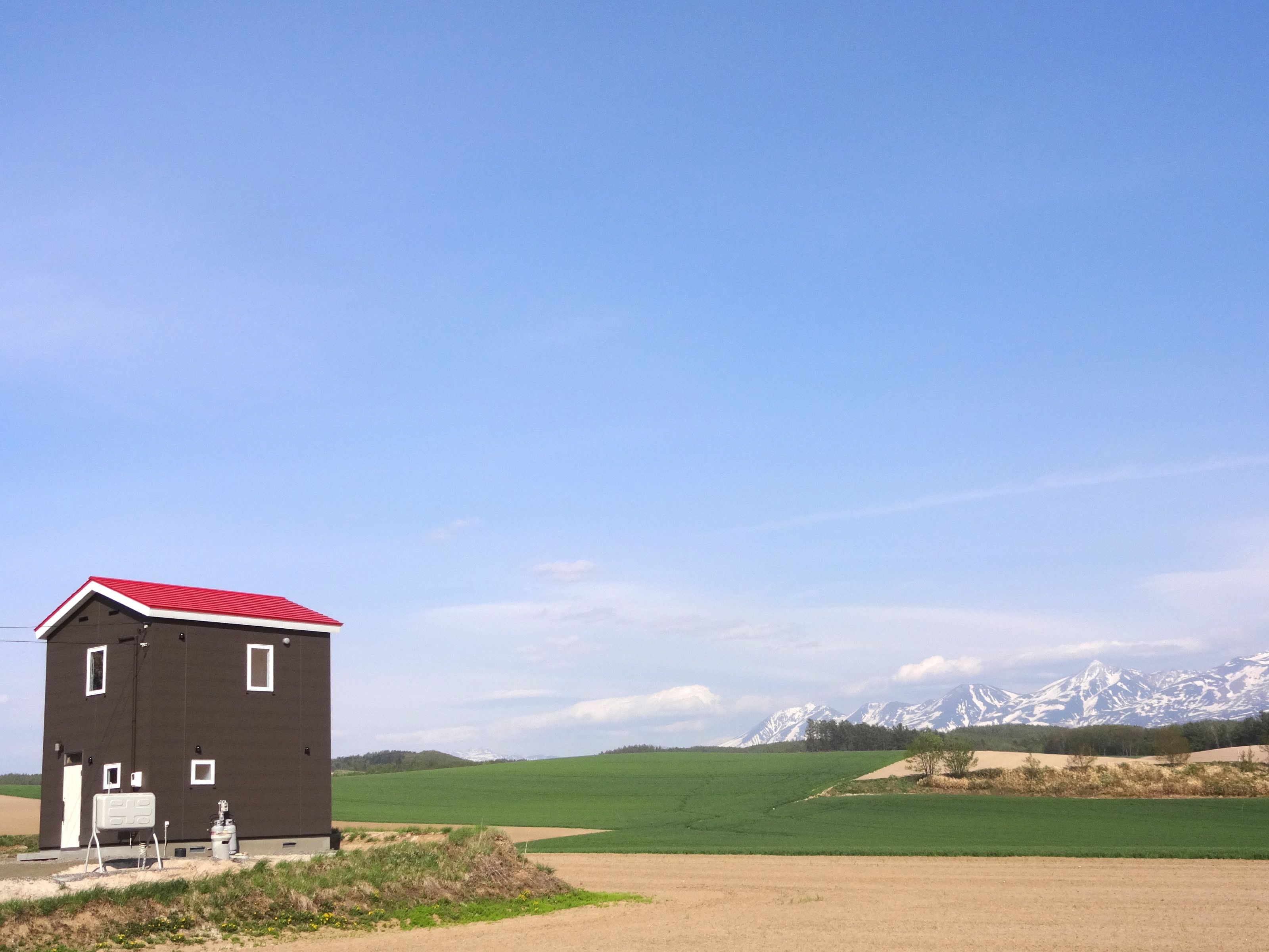 A small, dark building with a red roof, situated in a rural area with green fields and snow-capped mountains in the background.