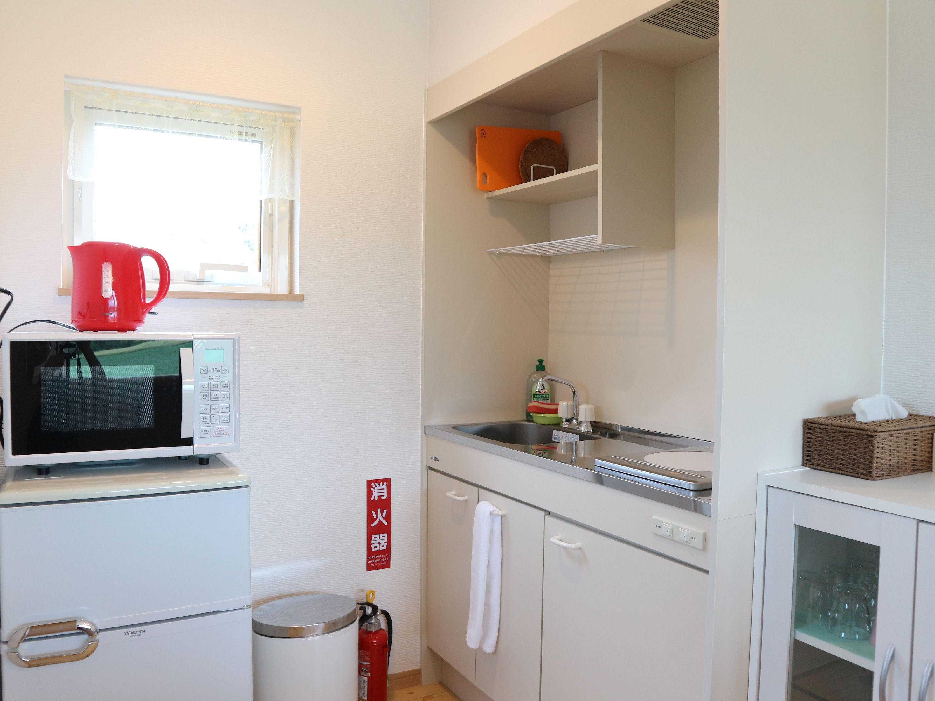 A compact kitchen with a microwave, a kettle, a sink, and a fire extinguisher.