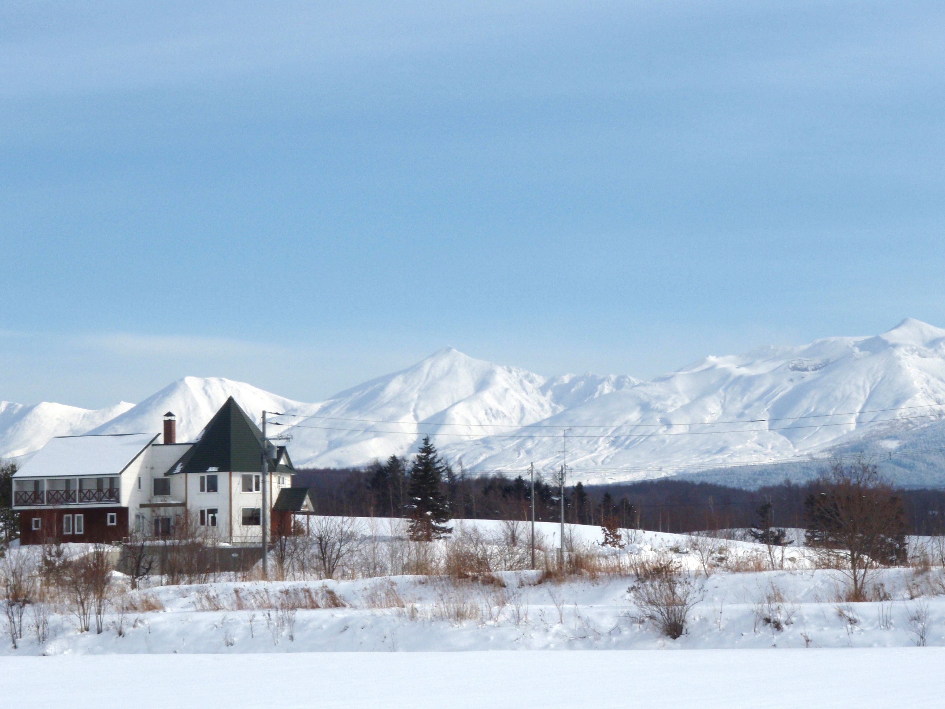 A large house with a green roof and white walls, surrounded by a snowy landscape and mountains in the background.