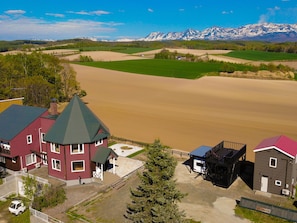 A two-story building with a red facade and a green roof, adjacent to a large open field and a mountain range in the background. - Pension Landscape Furano (Kamifurano)