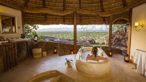 A spa room with a large bathtub, a wooden bench, and a view of the landscape. - Elewana Elsa’s Kopje (Meru National Park)