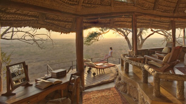 A high-ceilinged room with a thatched roof, a wooden bench, a table with books, and a view of the landscape. - Elewana Elsa’s Kopje (Meru National Park)