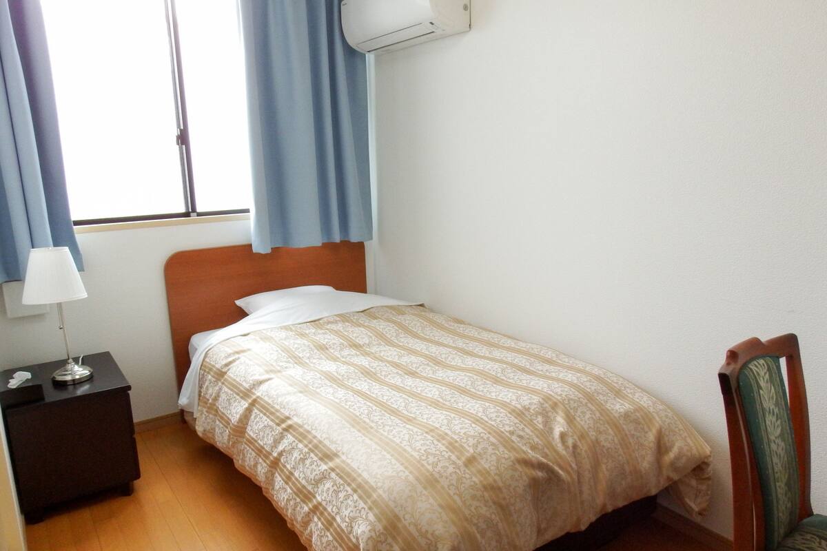 Habitación de hotel con una cama individual, una mesita de noche con lámpara, una silla de madera y un equipo de aire acondicionado en la pared.
