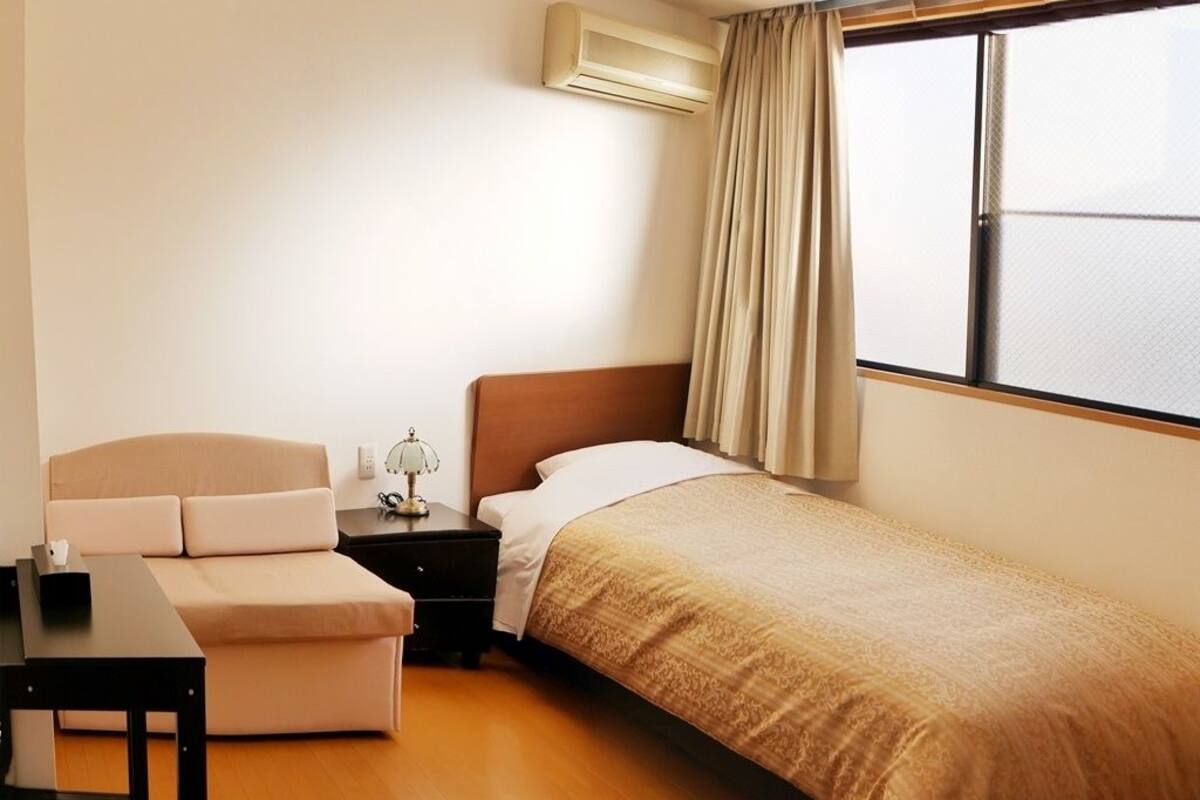 Una cama individual con una manta color beige, una mesita de noche con lámpara, un pequeño sofá y una ventana con cortinas.