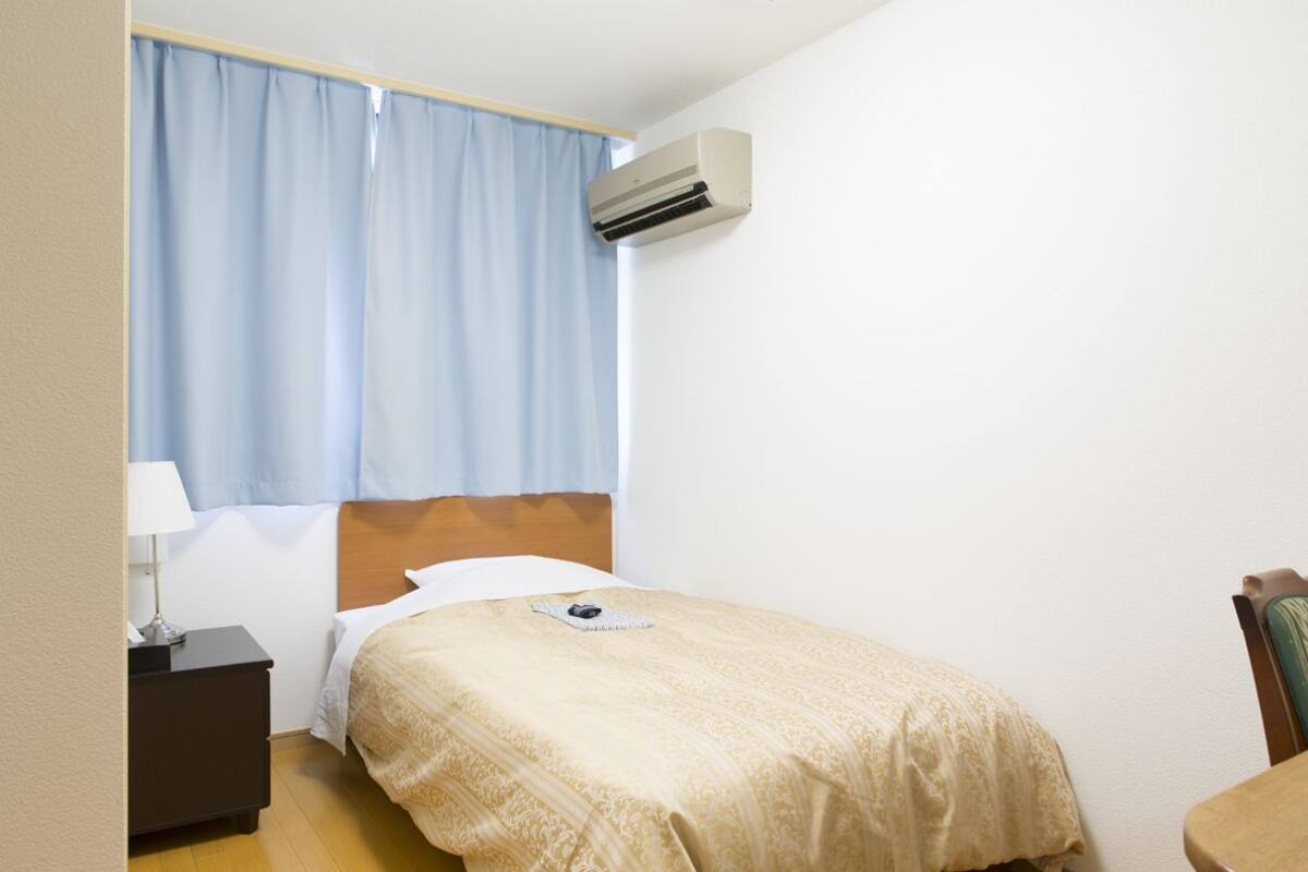 Una habitación de hotel con cama, mesita de noche, lámpara, equipo de aire acondicionado y una silla.
