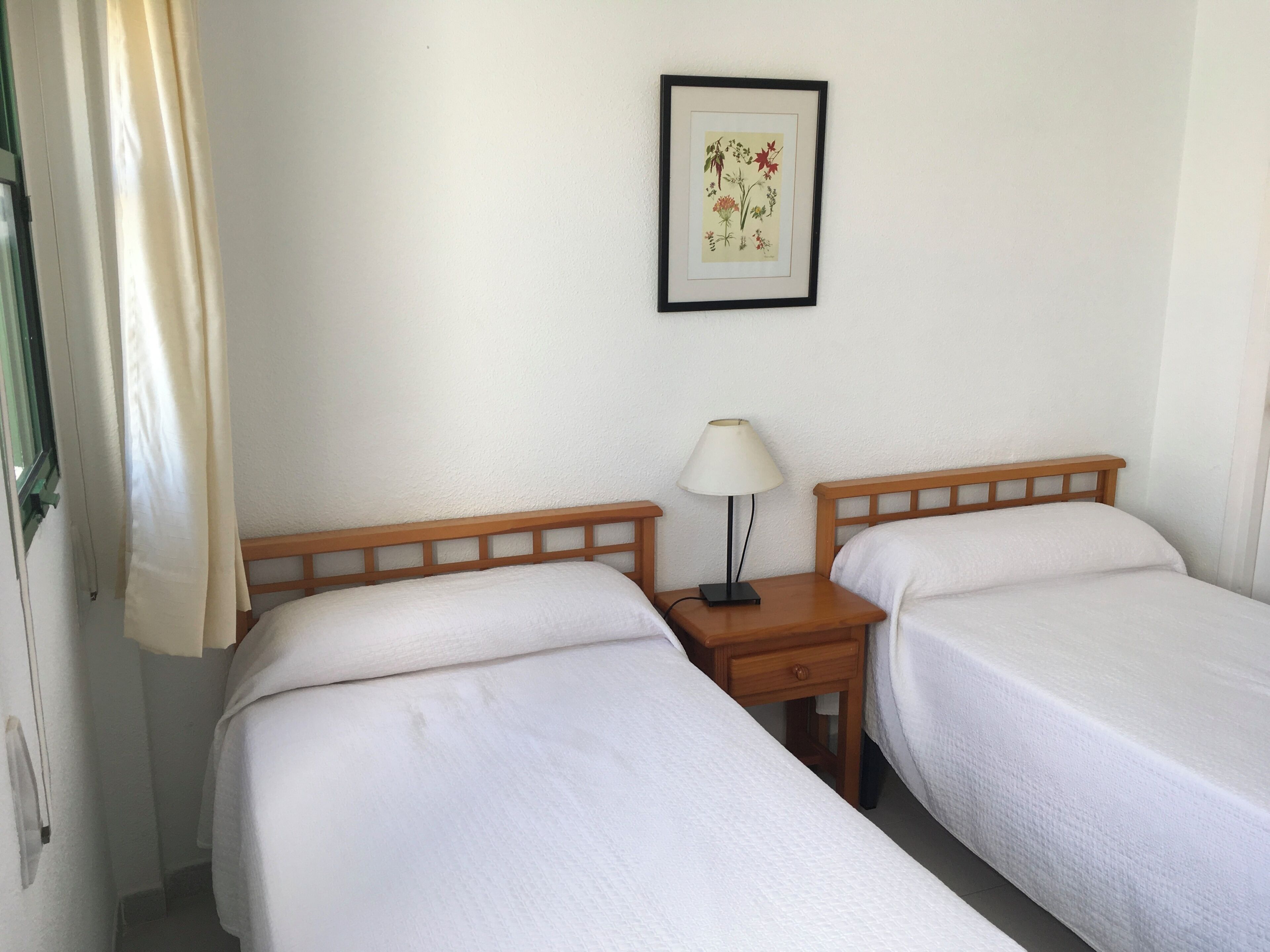 Een kamer met twee bedden, een nachtkastje met een lamp en een lijst met een botanische illustratie aan de muur.