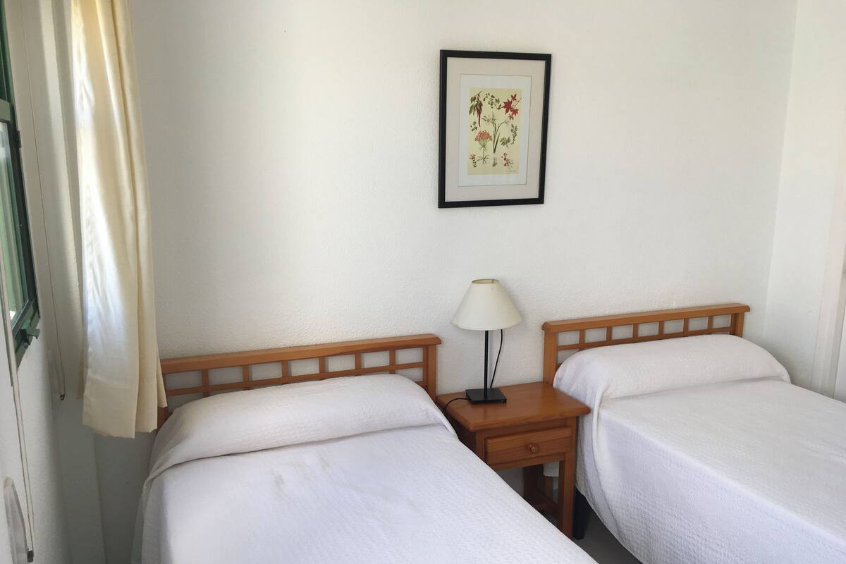 A room with two beds, a nightstand with a lamp, and a framed botanical illustration on the wall.