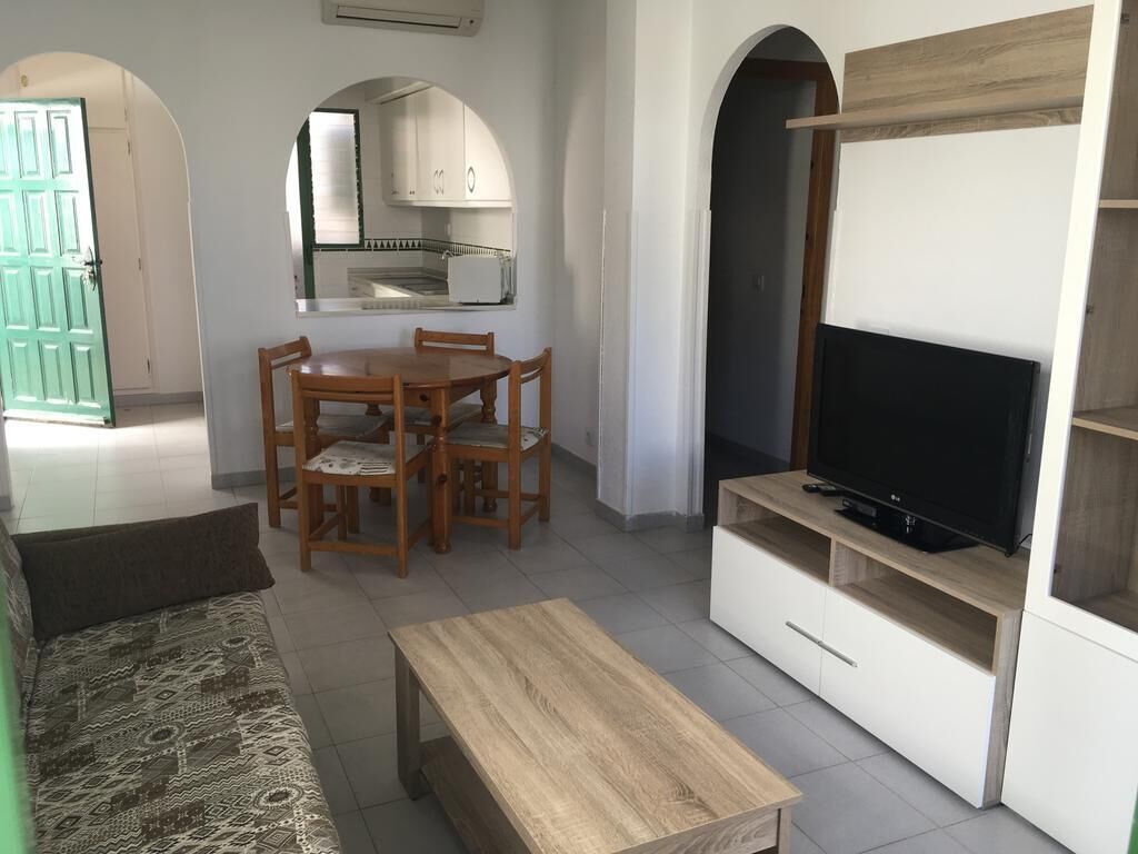 Een woonkamer met een bank, een houten salontafel, een tv op een standaard, een eettafel met stoelen en een keukenhoek met kasten en een raam.