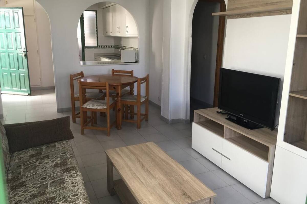 A living room with a sofa, a wooden coffee table, a TV on a stand, a dining table with chairs, and a kitchen area with cabinets and a window.