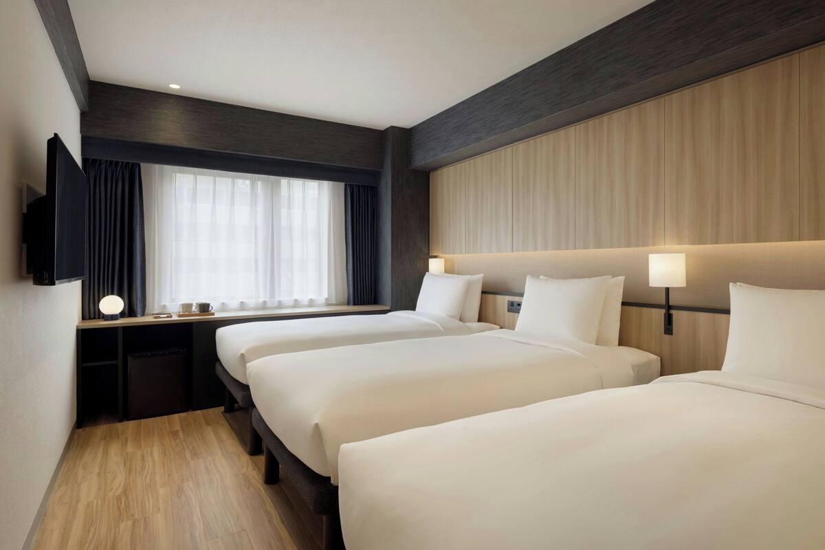 Habitación de hotel con dos camas, televisión, escritorio con lámpara y ventana con cortinas.