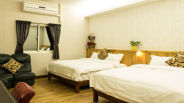 Habitación de hotel con dos camas, cabecero de madera, ventana con cortinas, sillón de cuero y una pequeña mesa.