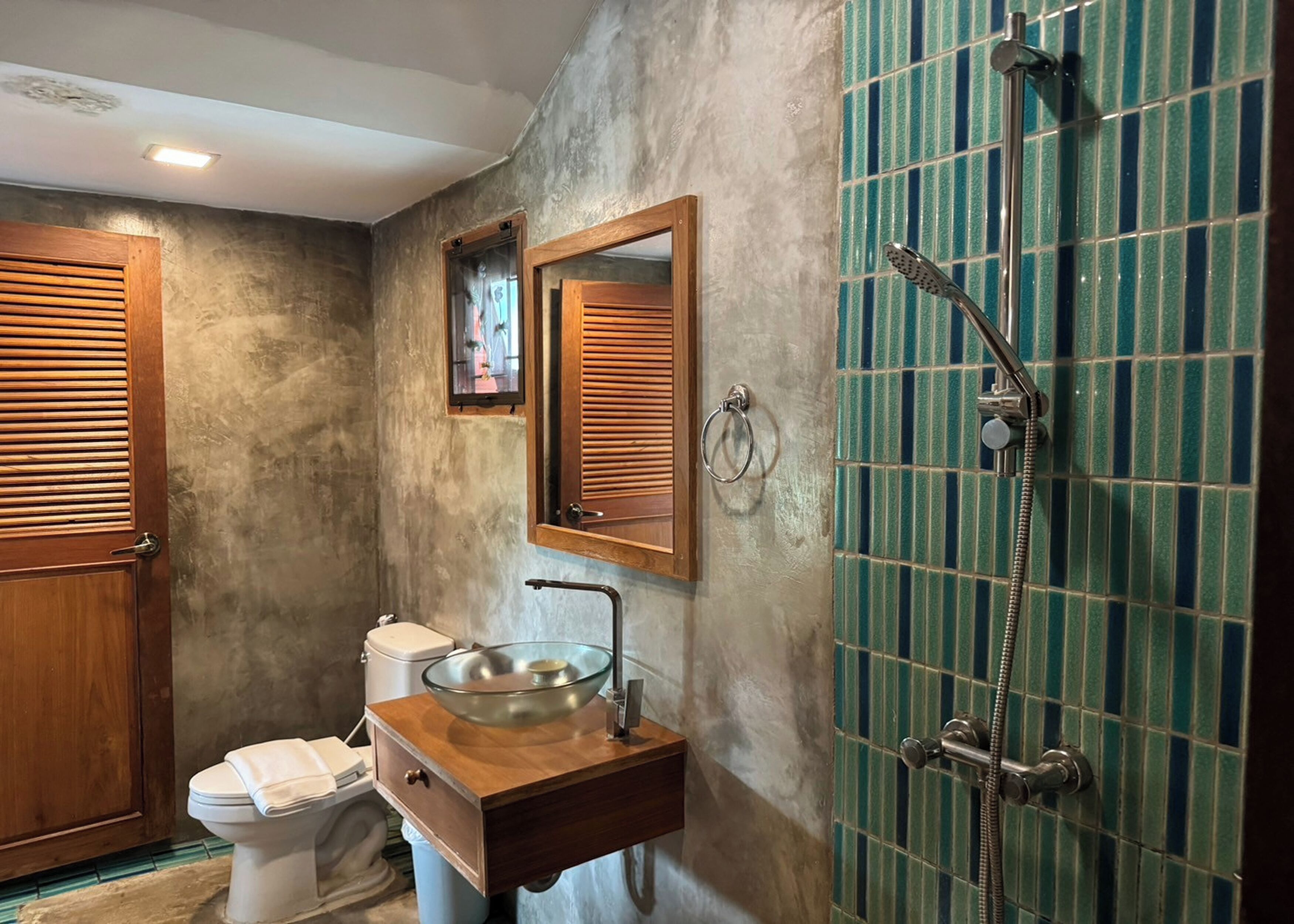 A bathroom with a wooden vanity, a glass sink, a toilet, and a shower with green tiles.