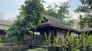 Een rustieke houten hut met een veranda, omgeven door weelderig groen en een klein vijvertje.