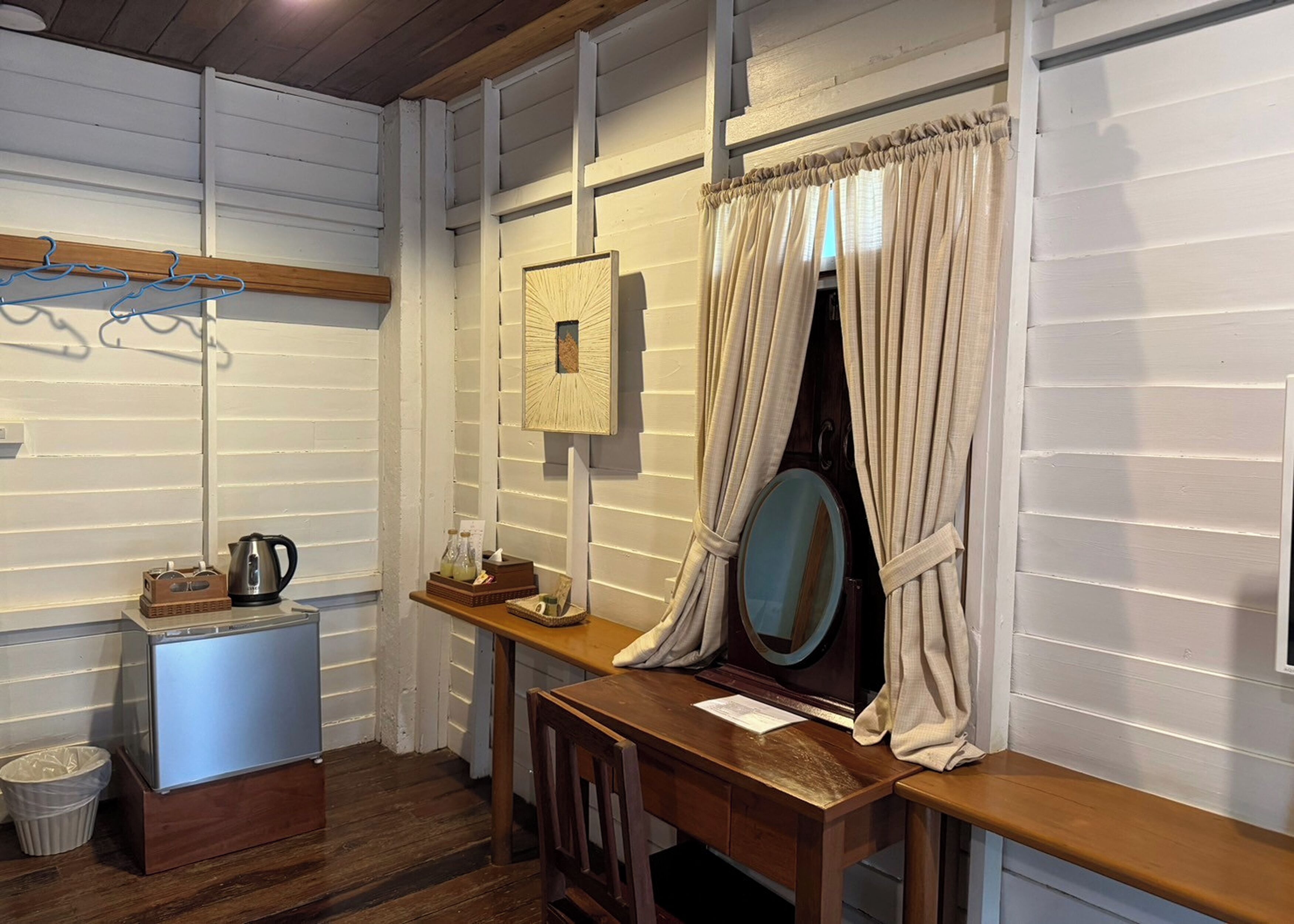 Quarto compacto com um banco de madeira, uma mesinha com uma chaleira, um espelho e uma janela com cortinas.