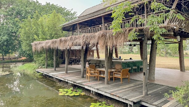 Een houten terras met zitplaatsen buiten en een rieten dakconstructie boven een vijver.